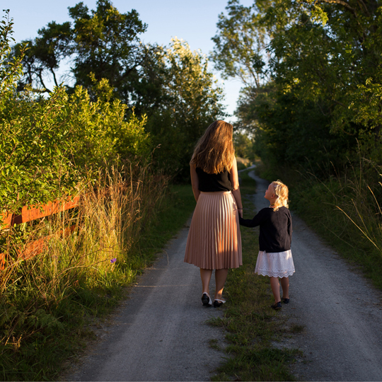 SOLOMÜTTER Info-Kachel: Bild, Alleinerziehende Mutter mit Tochter, Hand in Hand, spazierengehend auf einem Waldweg in untergehender Sonne